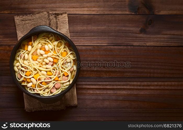 Chilean traditional Porotos con Riendas (beans with reins) dish of cooked dried beans with pumpkin, onion, spaghetti and sausage, served in rustic bowl, photographed overhead on dark wood with natural light