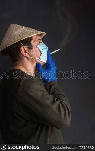 Chinese adult man in traditional Asian conical straw hat smokes a cigarette in a virus-protective medical mask on a dark background