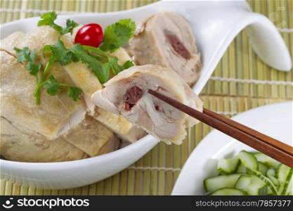 Chinese sliced roasted chicken, with chopsticks holding a single piece, in white bowl with sliced cucumber as side dish under natural bamboo place mat