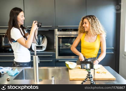 Chinese woman taping Caucasian woman cutting melon in a kitchen.