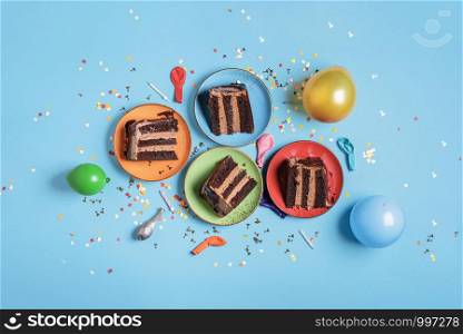 Chocolate birthday cake slices on plates, candles, balloons, and confetti on a blue table. Above view of festive dessert. New year celebration concept