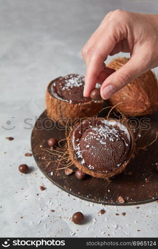 Chocolate brown homemade dessert in a coconut shell with girls hand decorating ice cream on a gray concrete background with place for text. Summer dessert for vegetarian.. A woman decorates a homemade chocolate ice cream in a coconut with chocolate balls on a board on a gray stone background.
