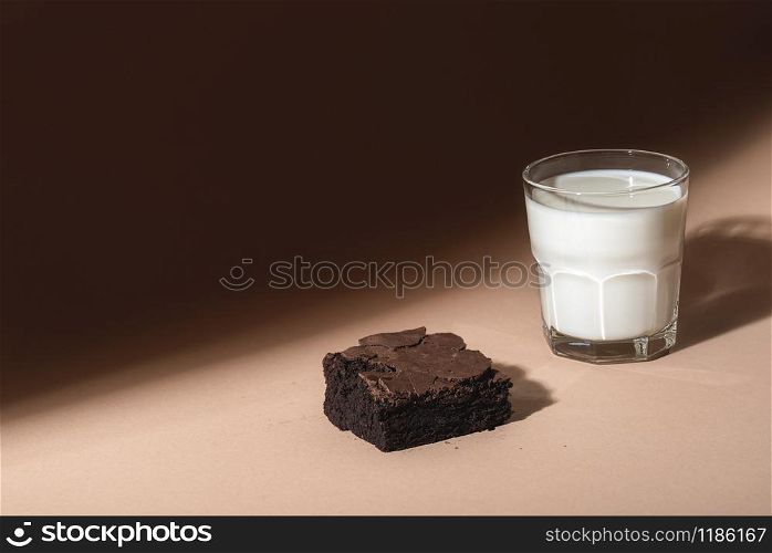 Chocolate brownie cake and a glass of warm milk on a brown background, in sunlight. Indulgence food. Chocolate dessert and milk. Unhealthy breakfast.