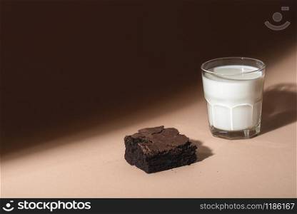 Chocolate brownie cake and a glass of warm milk on a brown background, in sunlight. Indulgence food. Chocolate dessert and milk. Unhealthy breakfast.