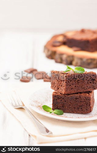 Chocolate brownie square pieces in stack on white plate decorated with mint leaves and cocoa powder on white vintage wooden background. American traditional delicious dessert. Close up photography.. Chocolate brownie square pieces in stack on white plate decorated with mint leaves and cocoa powder on white vintage wooden background. American traditional delicious dessert. Close up photography