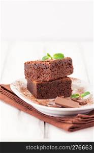Chocolate brownie square pieces in stack on white plate decorated with mint leaves and cocoa powder on white vintage wooden background. American traditional delicious dessert. Close up photography.. Chocolate brownie square pieces in stack on white plate decorated with mint leaves and cocoa powder on white vintage wooden background. American traditional delicious dessert. Close up photography