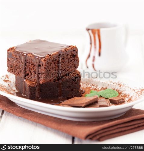 Chocolate brownie square pieces in stack on white plate decorated with mint leaves and cocoa powder on white vintage wooden background. American traditional delicious dessert. Close up photography.. Chocolate brownie square pieces in stack on white plate decorated with mint leaves and cocoa powder on white vintage wooden background. American traditional delicious dessert. Close up photography