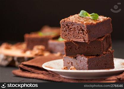 Chocolate brownie square pieces in stack on white plate with walnuts, decorated with mint leaves and cocoa on black background. Delicious dessert. Dark mood. Close up photography. Selective focus.. Chocolate brownie square pieces in stack on white plate with walnuts, decorated with mint leaves and cocoa on black background. Delicious dessert. Dark mood. Close up photography. Selective focus