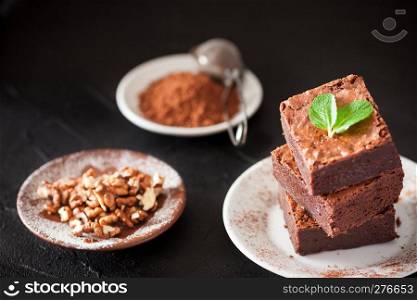 Chocolate brownie square pieces in stack on white plate with walnuts, decorated with mint leaves and cocoa on black background. Delicious dessert. Dark mood. Close up photography. Selective focus.. Chocolate brownie square pieces in stack on white plate with walnuts, decorated with mint leaves and cocoa on black background. Delicious dessert. Dark mood. Close up photography. Selective focus