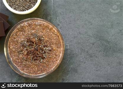Chocolate chia seed pudding in glass bowl with chia seeds and chocolate shavings on top, photographed overhead on slate with natural light