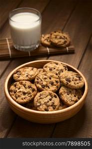 Chocolate chip cookies in wooden bowl with a glass of cold milk in the back, photographed on wood with natural light (Selective Focus, Focus on the cookie in the middle)