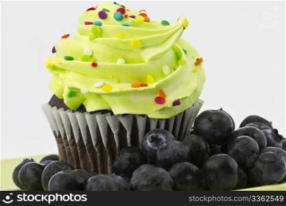 Chocolate cupcake with green frosting is surrounded by summer sweet blueberries