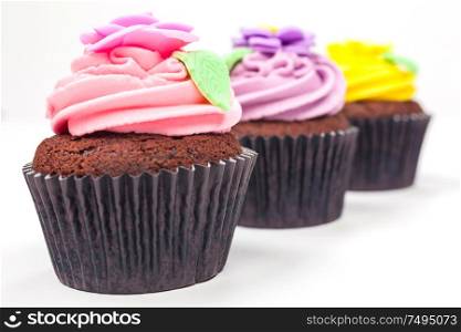 Chocolate cupcakes or cup cakes with icing or frosting, pink, purple, yellow with green leaves, rose and floral decorations photographed on a white background