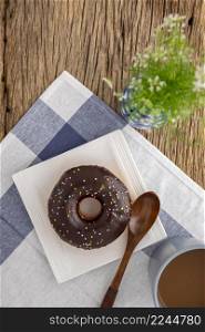 chocolate donut and wooden spoon in white square ceramic plate beside coffee and vase on napkin and rustic natural wood texture background, easy meal for break time, top view