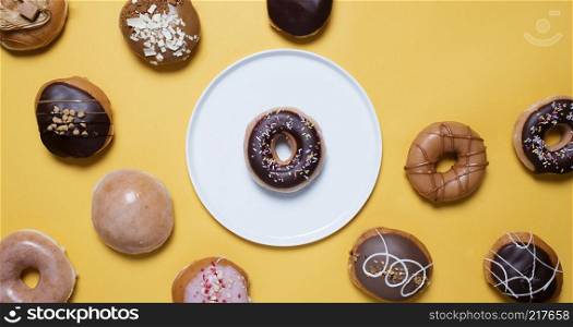 Chocolate donut with sprinkles on a white plate surrounded by various doughnuts on a yellow background