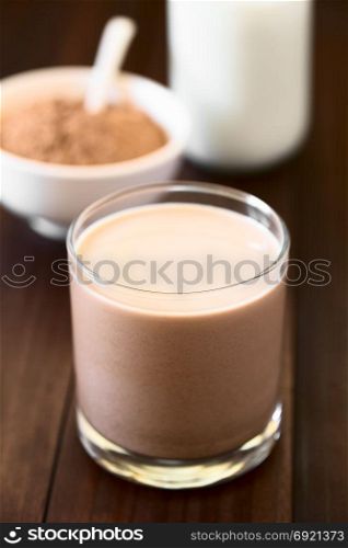 Chocolate milk drink in glass, chocolate or cocoa powder and milk in the back, photographed with natural light (Selective Focus, Focus on the front of the glass rim). Chocolate Milk Drink