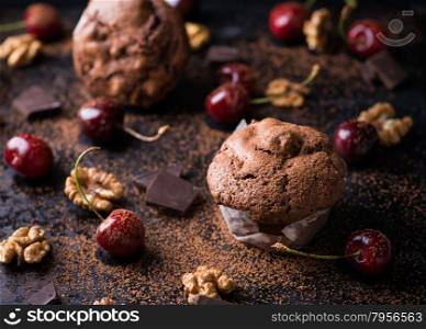 Chocolate muffins on dark background, cocoa powder, walnuts, cherries and chocolate chunks as decoration, selective focus