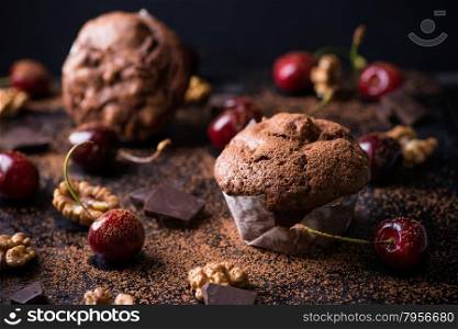Chocolate muffins on dark background, cocoa powder, walnuts, cherries and chocolate chunks as decoration, selective focus