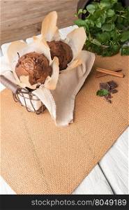 Chocolate muffins with chocolate slices in basket, cinnamom sticks and mint on placemat