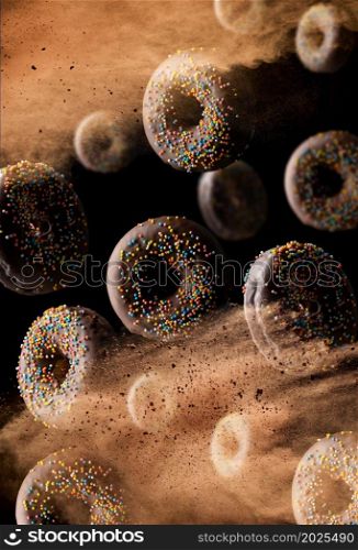 chocolate round donuts with multicolored sugar sprinkles levitate in a cloud of brown cocoa on a black background. Powder flies up