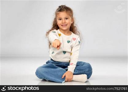 choice and childhood and people concept - beautiful smiling girl sitting on floor and pointing finger to camera over grey background. smiling girl pointing finger to camera