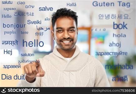 choice, gesture and people concept - smiling indian man pointing finger to camera at office over greeting words in different foreign languages. indian man over words in foreign languages