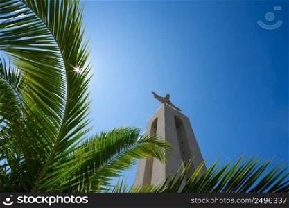 Christ the King Statue through green palm leaf at clear summer sunny day in Lisbon Portugal