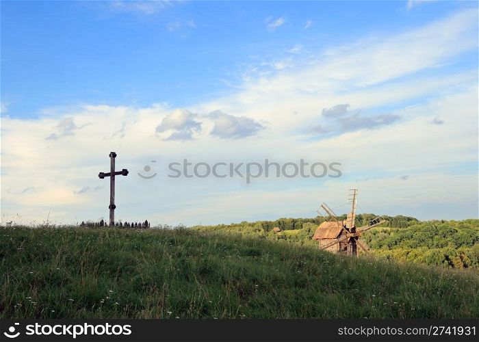Christian wood cross on grass overgrown hill (forest colony and windmill beyond)