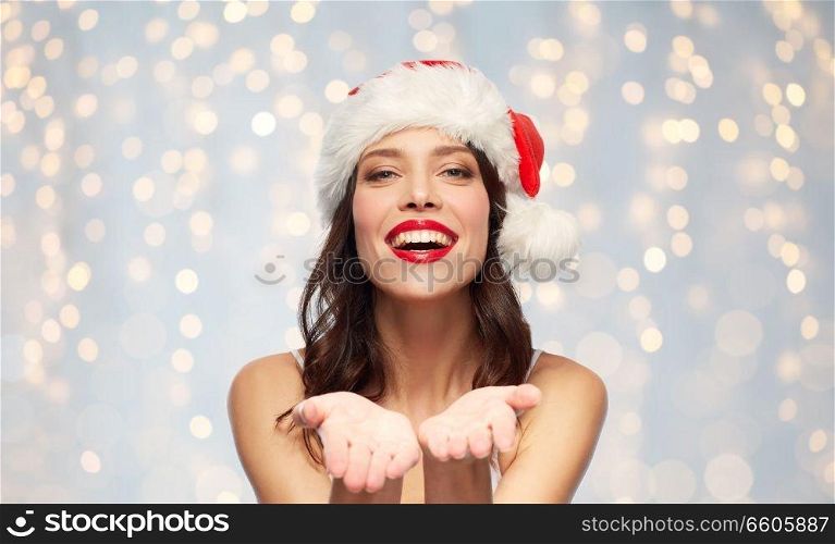 christmas and holidays concept - happy smiling young woman with empty hands in santa hat over festive lights background. woman with empty hands in santa hat on christmas