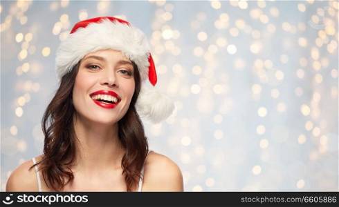 christmas and holidays concept - happy smiling young woman with red lipstick in santa hat over festive lights background. woman with red lipstick in santa hat on christmas