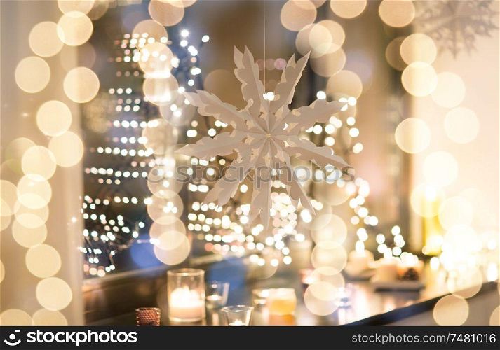 christmas and holidays concept - paper snowflake decoration hanging over garland lights and candles at night window. paper snowflake decoration hanging on window
