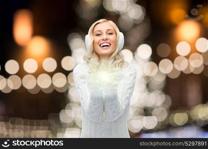 christmas and magic concept - smiling young woman in earmuffs and sweater holding fairy dust over night lights background. happy woman over christmas lights