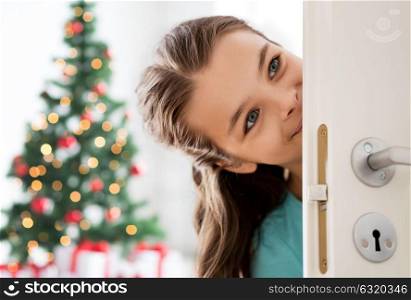christmas and people concept - happy smiling beautiful girl behind door at home. happy smiling beautiful girl behind door at home