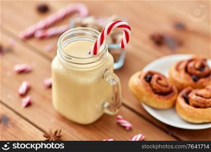 christmas and seasonal drinks concept - eggnog in glass mug with candy cane decoration, cinnamon buns and aromatic spices on wooden background. eggnog with candy cane in mug and cinnamon buns
