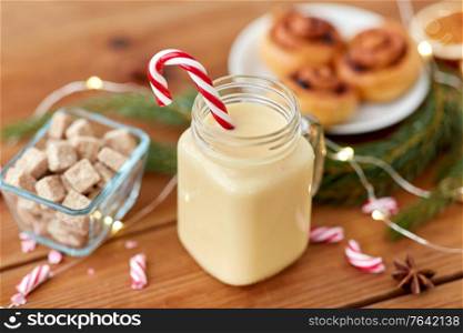 christmas and seasonal drinks concept - eggnog in glass mug with candy cane decoration, cinnamon buns, fir tree brunch and garland lights on wooden background. eggnog with candy cane in mug and cinnamon buns