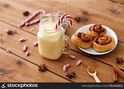christmas and seasonal drinks concept - eggnog in glass mug with candy cane decoration, cinnamon buns and aromatic spices on wooden background. eggnog with candy cane in mug and cinnamon buns