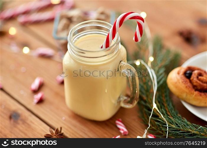 christmas and seasonal drinks concept - eggnog in glass mug with candy cane decoration, cinnamon buns, fir tree brunch and garland lights on wooden background. eggnog with candy cane in mug and cinnamon buns