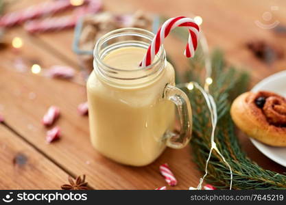 christmas and seasonal drinks concept - eggnog in glass mug with candy cane decoration, cinnamon buns, fir tree brunch and garland lights on wooden background. eggnog with candy cane in mug and cinnamon buns