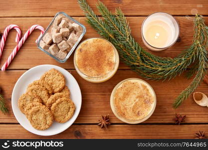 christmas and seasonal drinks concept - glasses of eggnog with oatmeal cookies, candy canes, sugar, fir tree branches and candle burning on wooden background. glasses of eggnog, oatmeal cookies and fir branch