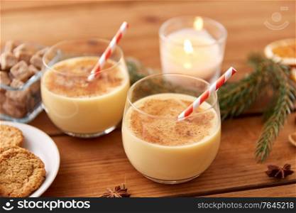 christmas and seasonal drinks concept - glasses of eggnog with oatmeal cookies, candy canes, sugar, fir tree branches and candle burning on wooden background. glasses of eggnog, oatmeal cookies and fir branch
