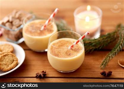 christmas and seasonal drinks concept - glasses of eggnog with oatmeal cookies, candy canes, sugar, fir tree branches and candle burning on wooden background. glasses of eggnog, oatmeal cookies and fir branch