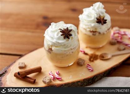 christmas and seasonal drinks concept - glasses of eggnog with whipped cream topping, candy canes, sugar and cinnamon on wood cut board. glasses of eggnog with whipped cream and anise