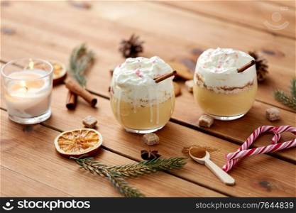 christmas and seasonal drinks concept - glasses of eggnog with whipped cream topping and cinnamon, fir tree branches, brown sugar and candle burning on wooden background. glasses of eggnog with whipped cream and spices