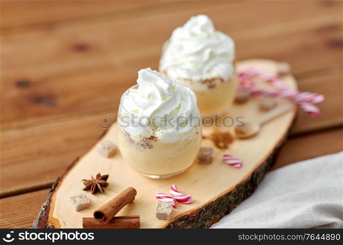 christmas and seasonal drinks concept - glasses of eggnog with whipped cream topping, candy canes, sugar and cinnamon on wood cut board. glasses of eggnog with whipped cream and anise