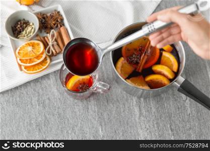 christmas and seasonal drinks concept - hand with ladle pouring hot mulled wine with orange slices and aromatic spices to glass cup on grey background. hand with ladle pouring hot mulled wine to glass