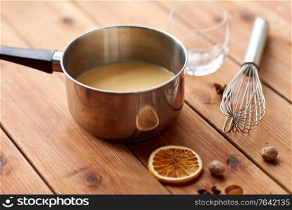christmas and seasonal drinks concept - pot with eggnog, whisk, glass and aromatic spices on wooden background. pot with eggnog, whisk and spices on wood