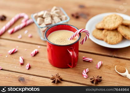 christmas and seasonal drinks concept - red cup of eggnog with candy cane, oatmeal cookies, fir tree branches and sugar on wooden background. cup of eggnog with candy cane, cookies and sugar