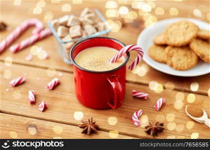 christmas and seasonal drinks concept - red cup of eggnog with candy cane, oatmeal cookies, fir tree branches and sugar on wooden background over lights. cup of eggnog with candy cane, cookies and sugar