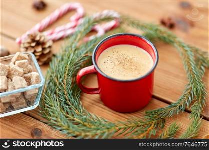 christmas and seasonal drinks concept - red cup of eggnog with cinnamon, fir tree branches, gingerbread, sugar and candy canes on wooden background. cup of eggnog, fir branches, gingerbread and sugar