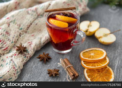 christmas and seasonal treats concept - glass of hot mulled wine with dry orange slicess and cinnamon, star anise, knitted scarf and apples on grey stone surface. glass of hot mulled wine with orange and cinnamon